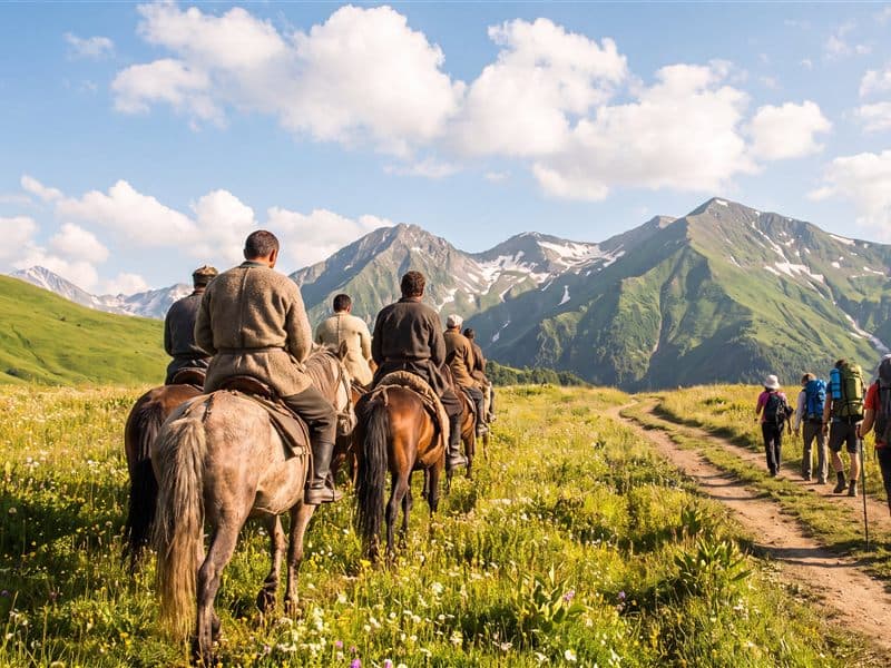 Riders on horseback crossing a wildflower meadow below green Caucasus peaks